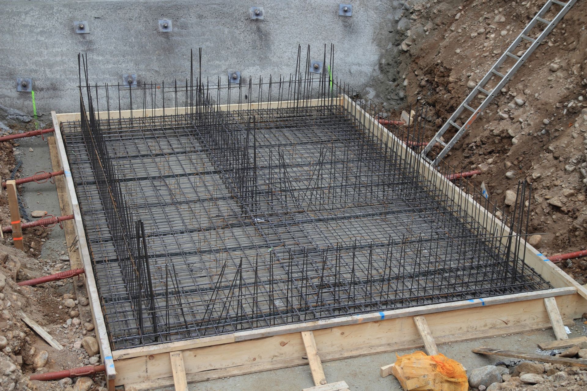 A construction site in West Palm Beach features a rectangular foundation being prepared. Steel rebar is arranged in a grid pattern within a wooden frame on the concrete slab. There is a dirt embankment around the site and a ladder leaning against the dirt on the right side.