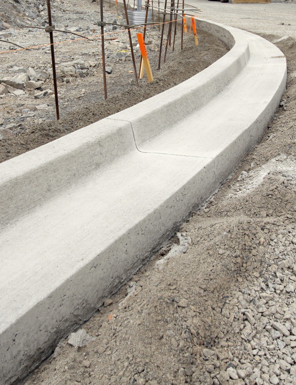 An under-construction site in Palm Beach County features a newly laid concrete curb with a smooth, rounded edge. The surrounding ground is covered with loose gravel and rocks. Rebar sticks out vertically behind the curb, indicating ongoing construction work involving decorative concrete designs.