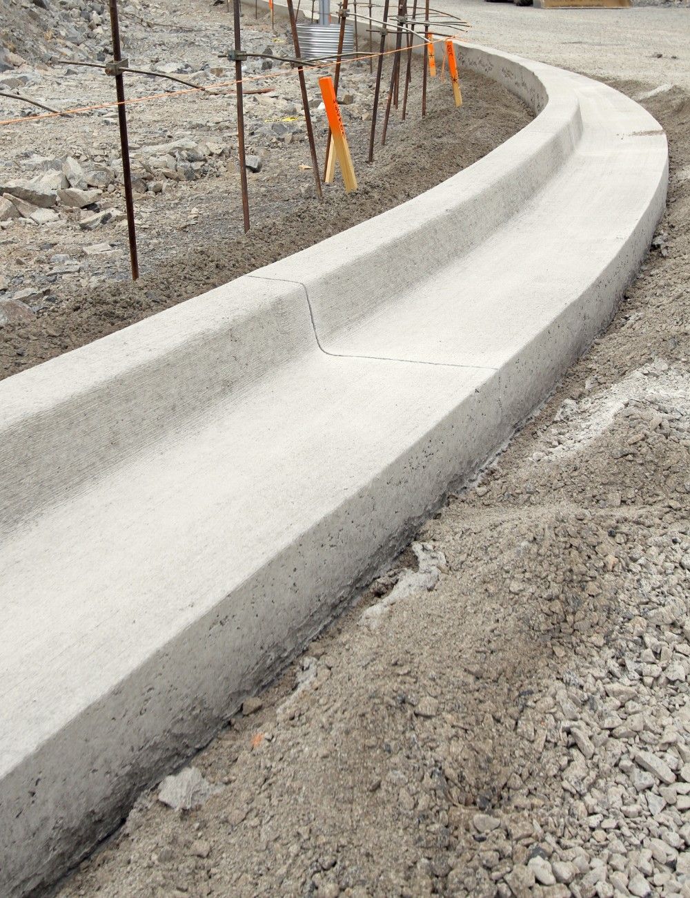 A freshly constructed decorative concrete curb curves smoothly along a dirt path. Wooden stakes with orange flags line the path on the opposite side of the curb, indicating an ongoing construction project. The surrounding area is cluttered with gravel and construction materials.