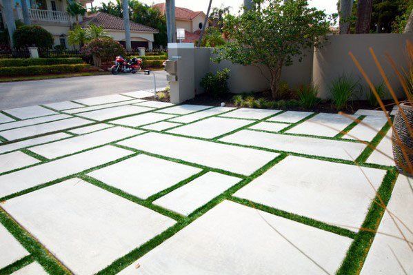 A modern driveway, showcasing decorative concrete slabs arranged in a geometric pattern with green grass filling the gaps, serves as a testament to expert driveway contracting in West Palm Beach. In the background, a residential neighborhood street features houses, trees, and a parked motorcycle.