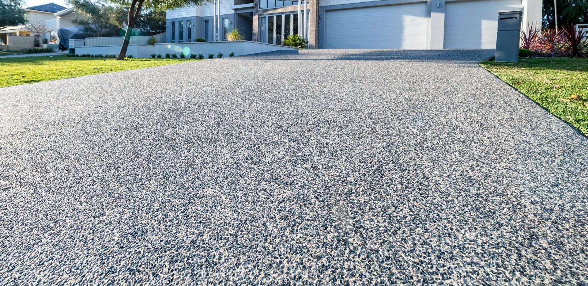 A smooth, expansive gray driveway leads up to a modern house with large windows and a garage. Expertly crafted by top Concrete Contractors in West Palm Beach FL, the driveway is flanked by green grass and a small landscaped area on one side. The scene is well-lit with the sun shining brightly.