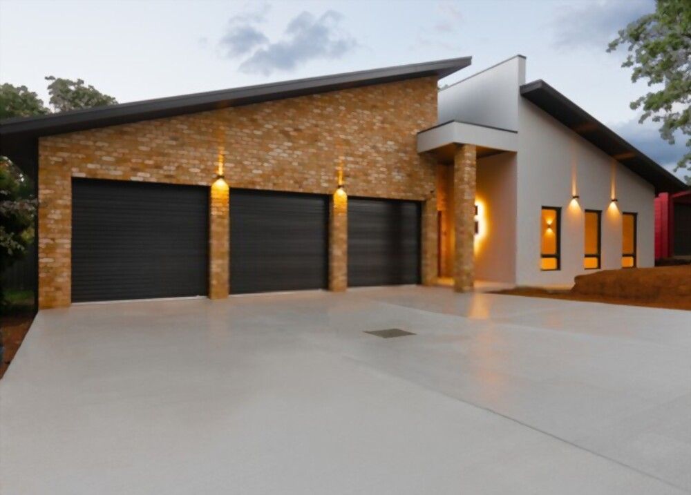 A modern house with a unique slanted roof design features a clean concrete driveway, expertly crafted by Concrete Contractor Juno Beach, leading to four black garage doors with illuminated brick accents. The exterior walls are a combination of brick and light-hued siding. The sky is partly cloudy.