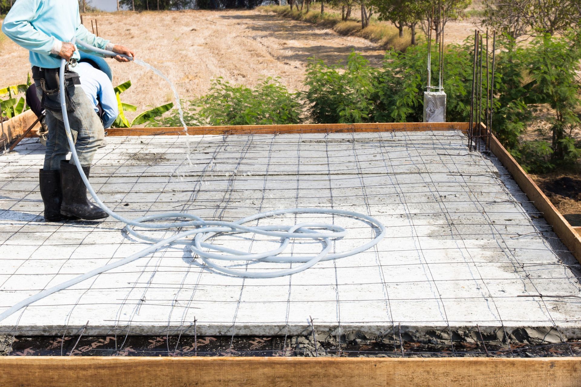 A worker in boots and a blue shirt sprays water onto a freshly poured concrete slab, essential for foundation contracting in West Palm Beach. The slab is framed with wooden boards and reinforced with steel mesh. Surrounded by dirt and greenery, the hose lies coiled on the concrete surface.