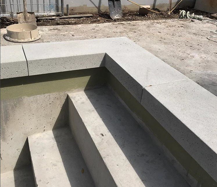 A construction site in Boynton Beach showcases newly poured concrete steps leading into an unfinished pool. The pool edges are covered with concrete coping, and building materials, such as buckets and tools, are scattered around the area—a typical scene for a stamped concrete contractor in Palm Beach County.