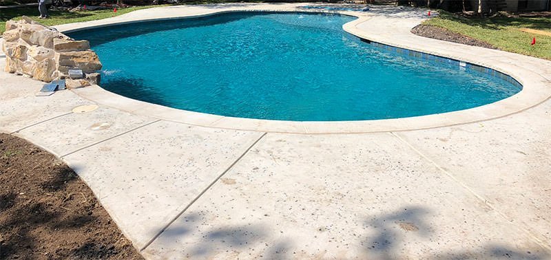 A backyard swimming pool with blue water and a surrounding stamped concrete deck crafted by experts in Palm Beach County. Decorative rocks are piled on one side of the freeform, curved pool, with lush greenery and trees in the background, creating a serene oasis in Delray Beach.