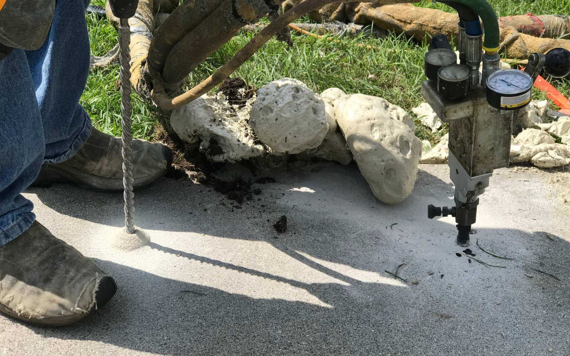 A person wearing jeans and work boots is using a drilling machine to inject materials into the concrete surface. The ground is partially covered with rocky residue and dirt. Green grass surrounds the work area, where various tools and equipment are scattered around—typical for a concrete repair project by a West Palm Beach FL restoration contractor.