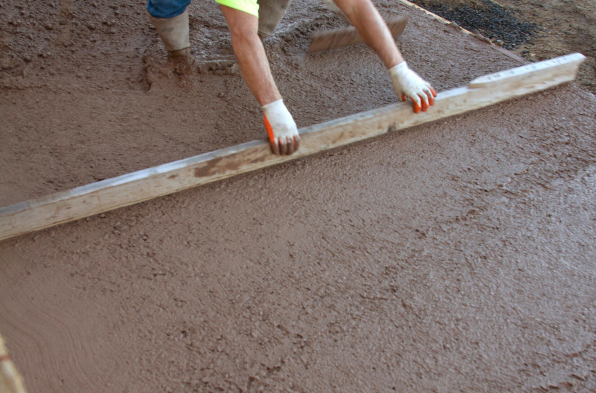 A person wearing gloves and boots is using a long wooden or metal tool to smooth and level a freshly poured Palm Beach stamped concrete surface. The concrete appears wet and is being spread evenly. The person, partially visible, focuses on the precise details of stamped concrete installation.