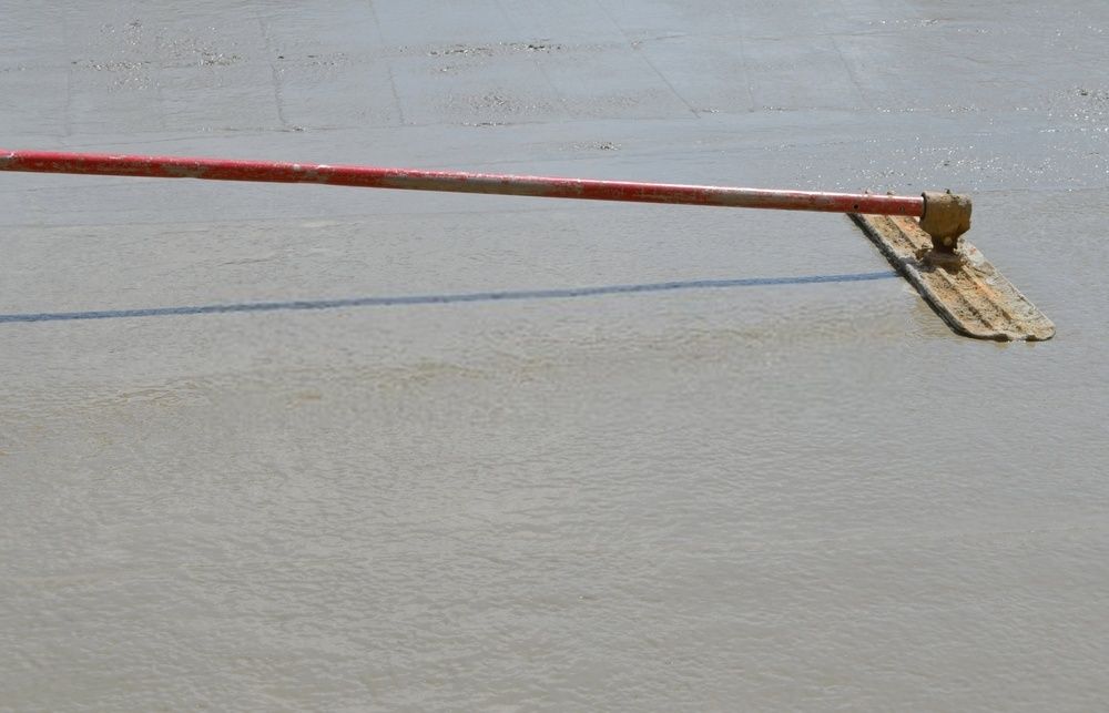 A close-up image of a concrete surface being smoothed using a long-handled float. The float, with its red handle, is in use by skilled concrete contractors to level and smooth the wet concrete. The surface is partially smoothed, showcasing different textures typical of decorative concrete services in Palm Beach County.