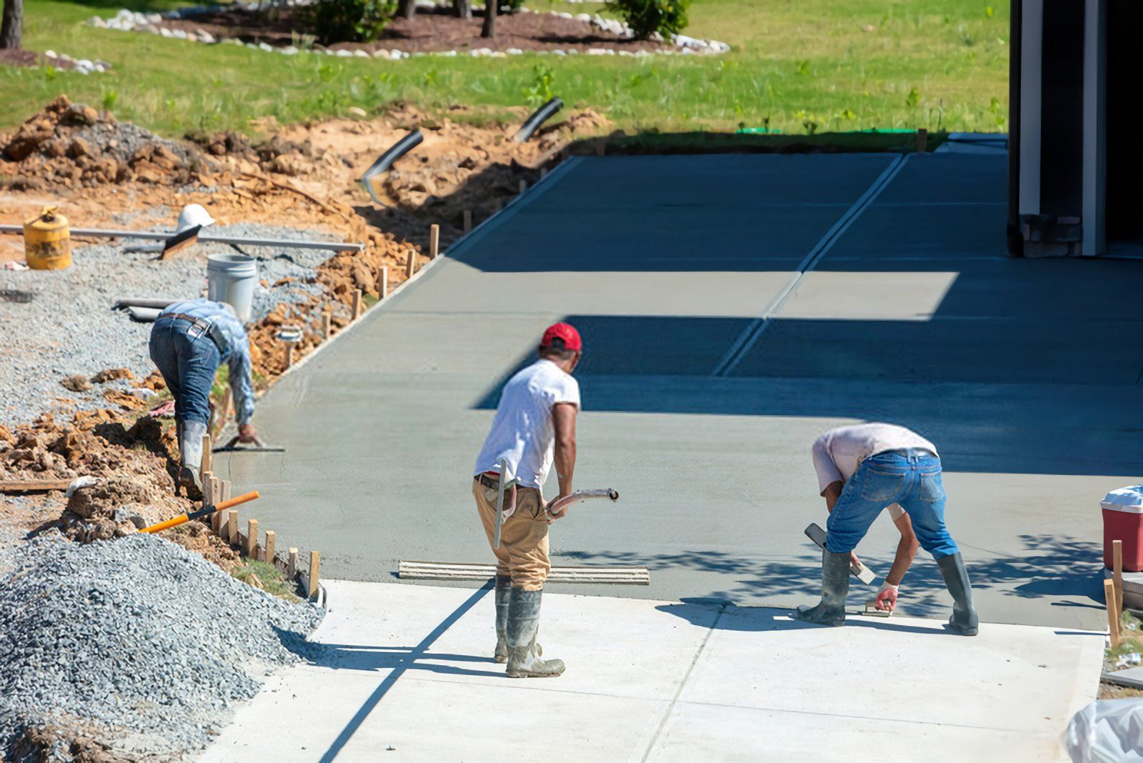 Three construction workers wearing casual clothing and boots are smoothing freshly poured decorative concrete on a driveway. The area around them, typical of West Palm Beach FL, is a mix of materials including gravel and dirt, with some equipment and tools visible. In the background, grass and a partially shaded area can be seen.