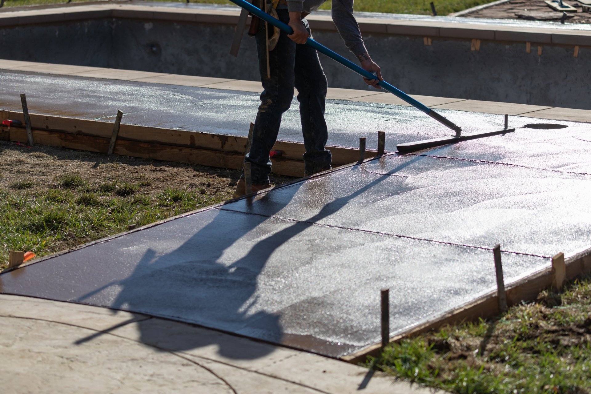 A construction worker smooths out wet, decorative concrete on a newly poured walkway next to an unfilled pool in Palm Beach County. The worker uses a long-handled tool and casts a pronounced shadow on the surface. Wooden forms and metal stakes outline the stamped concrete path.