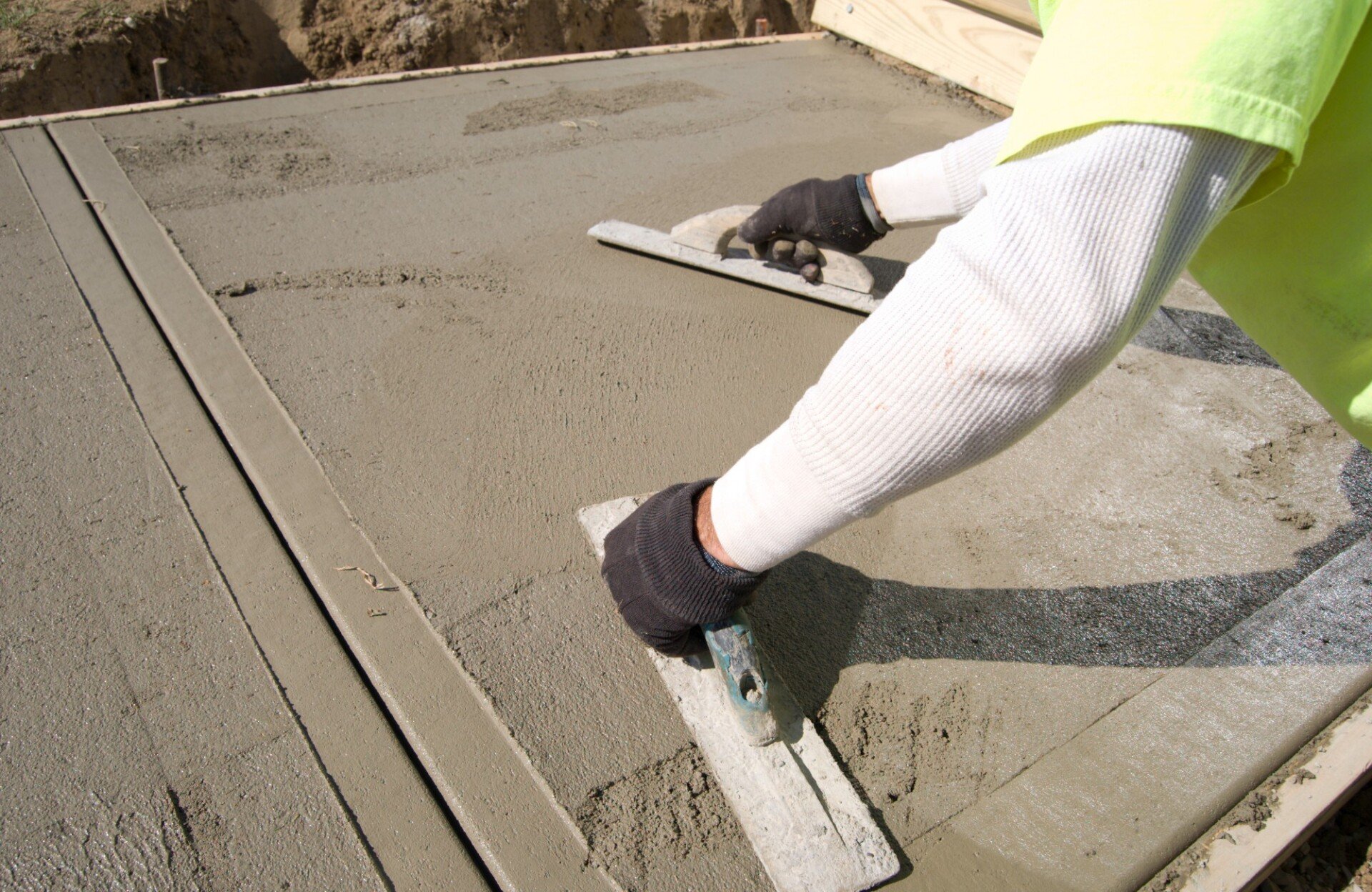 A worker, likely a stamped concrete contractor, wearing a long-sleeve shirt and gloves, smooths out wet cement on a flat surface using two trowels. The freshly poured concrete suggests an ongoing construction or paving project in Boynton Beach, Palm Beach County.