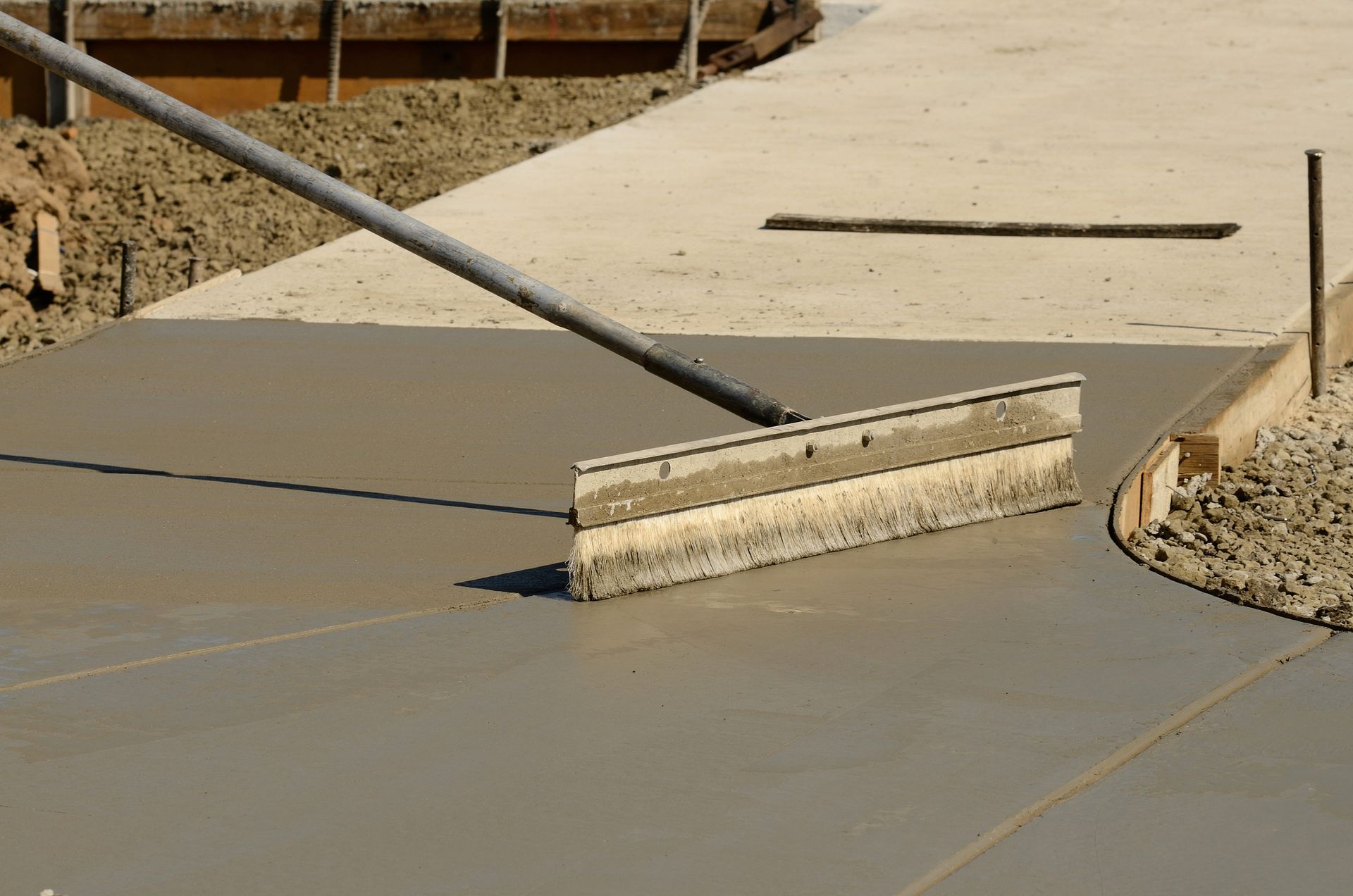 A concrete contractor in Palm City, FL is using a long-handled tool to smooth freshly poured concrete on a construction site. The wet concrete stretches out in a curved pathway, bordered by wooden forms, ready for stamped concrete. The surrounding area shows bare earth and construction materials.