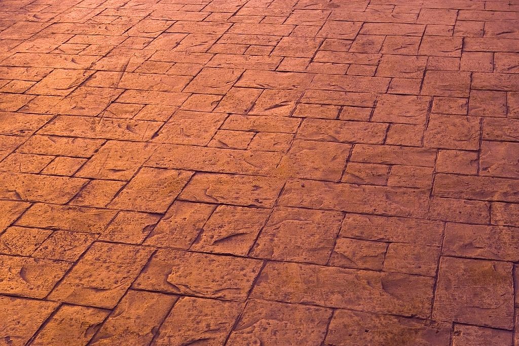 A close-up view of a ground surface with a patterned design made of textured, rectangular stone tiles in varying shades of warm brown. The sunlight accentuates the texture and color variations, creating a visually appealing effect that showcases the expertise of concrete contractors in Palm Beach County.