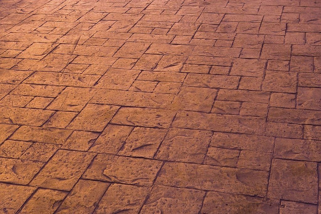 Image of a sunlit, brown, textured stamped concrete surface with a rectangular pattern. The stone-like texture and interlocking shapes cast subtle shadows and highlights. Perfect inspiration for your next project—contact our Concrete Contractors in West Palm Beach FL for Free Quotes.