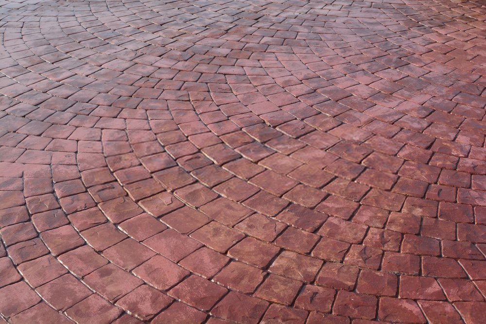 Close-up view of a circular paved surface made of red cobblestone bricks arranged in a repeating concentric pattern. The bricks have a slightly worn texture, creating a visually appealing geometric design, characteristic of expert work by Palm Beach County's top stamped concrete contractor.