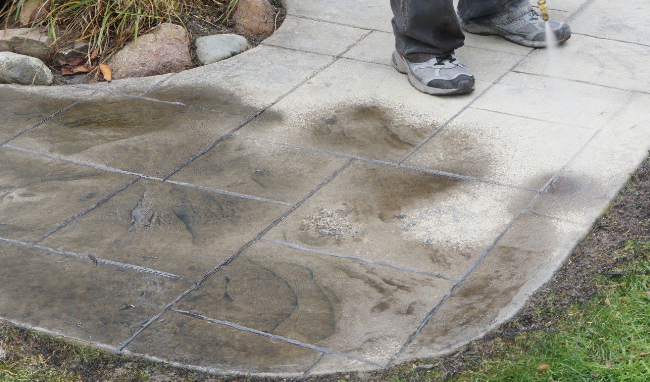 A person in Jupiter, FL, is pressure washing a patio with stone tiles. The cleaner sections appear significantly lighter, contrasting with the remaining dirty areas. The person's legs and sneakers are partially visible, along with surrounding grass and Palm Beach Stamped Concrete accents.
