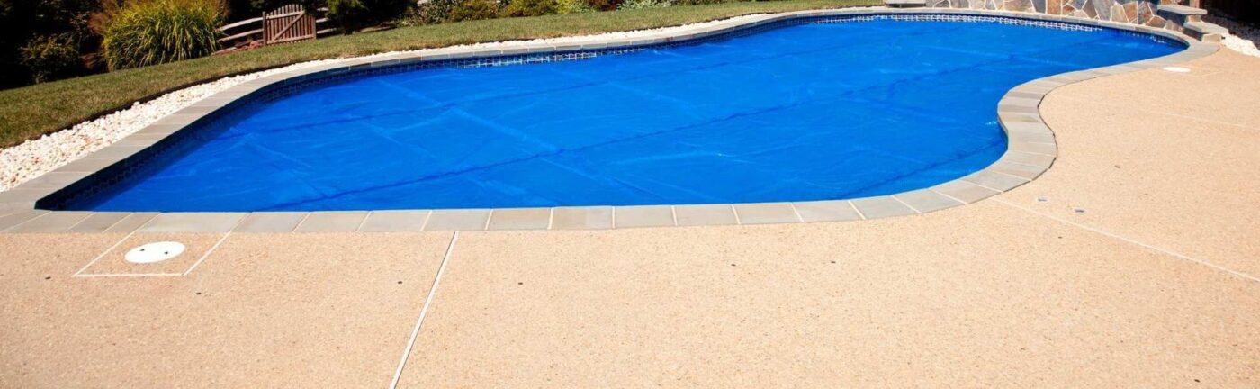 A curvy outdoor swimming pool covered with a blue pool cover, surrounded by a stone deck featuring cool decking, and a grassy area with landscaping in the background. The scene is brightly lit, suggesting a sunny day in West Palm Beach.