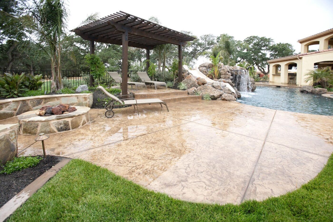 A luxurious backyard in West Palm Beach, FL features a stamped concrete patio crafted by expert concrete contractors, a pergola with lounge chairs, a built-in stone fire pit, and a pool with a natural-looking waterfall and rock formation. Lush landscaping surrounds the area, adding to the serene ambiance.