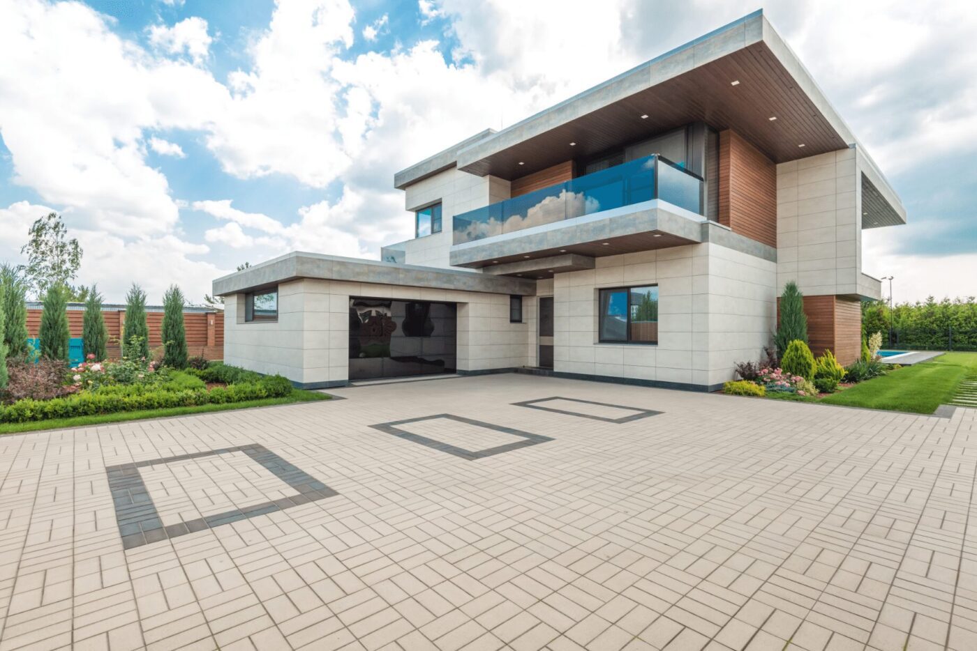 Modern two-story house with a flat roof, large windows, and a spacious driveway featuring geometric patterns crafted by top-notch driveway contracting. The home boasts a blend of white and wood paneling, a glass balcony, and is surrounded by landscaped greenery. Overhead, the sky is partly cloudy in West Palm Beach FL.