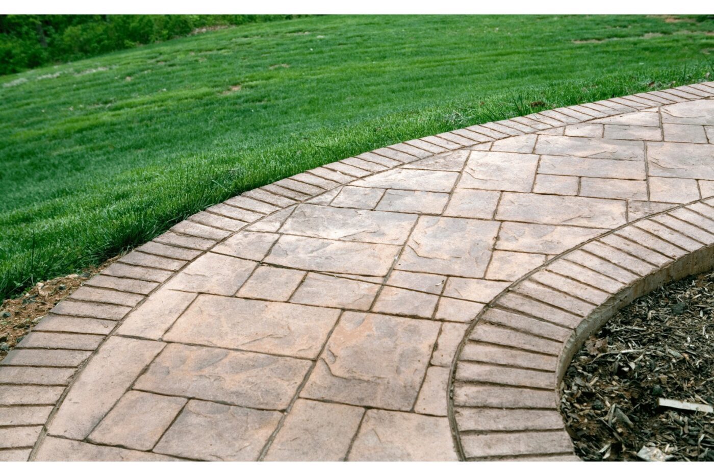 A curved stone walkway with a brick border leads through a neatly manicured green lawn in West Palm Beach, FL. The path consists of square and rectangular stones arranged in a pattern and edges a mulched garden bed, complementing the nearby concrete patio crafted by Palm Beach Stamped Concrete.